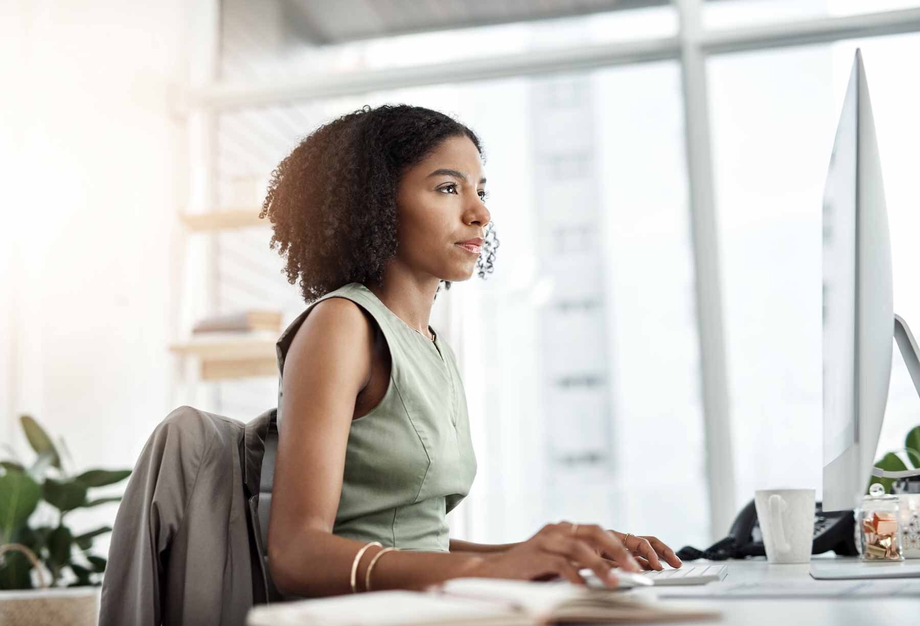 An African American woman with a focused expression is sitting at her desk, using a computer. She's on the HubSpot platform, analyzing the performance metrics of her latest blog posts displayed on the screen.
