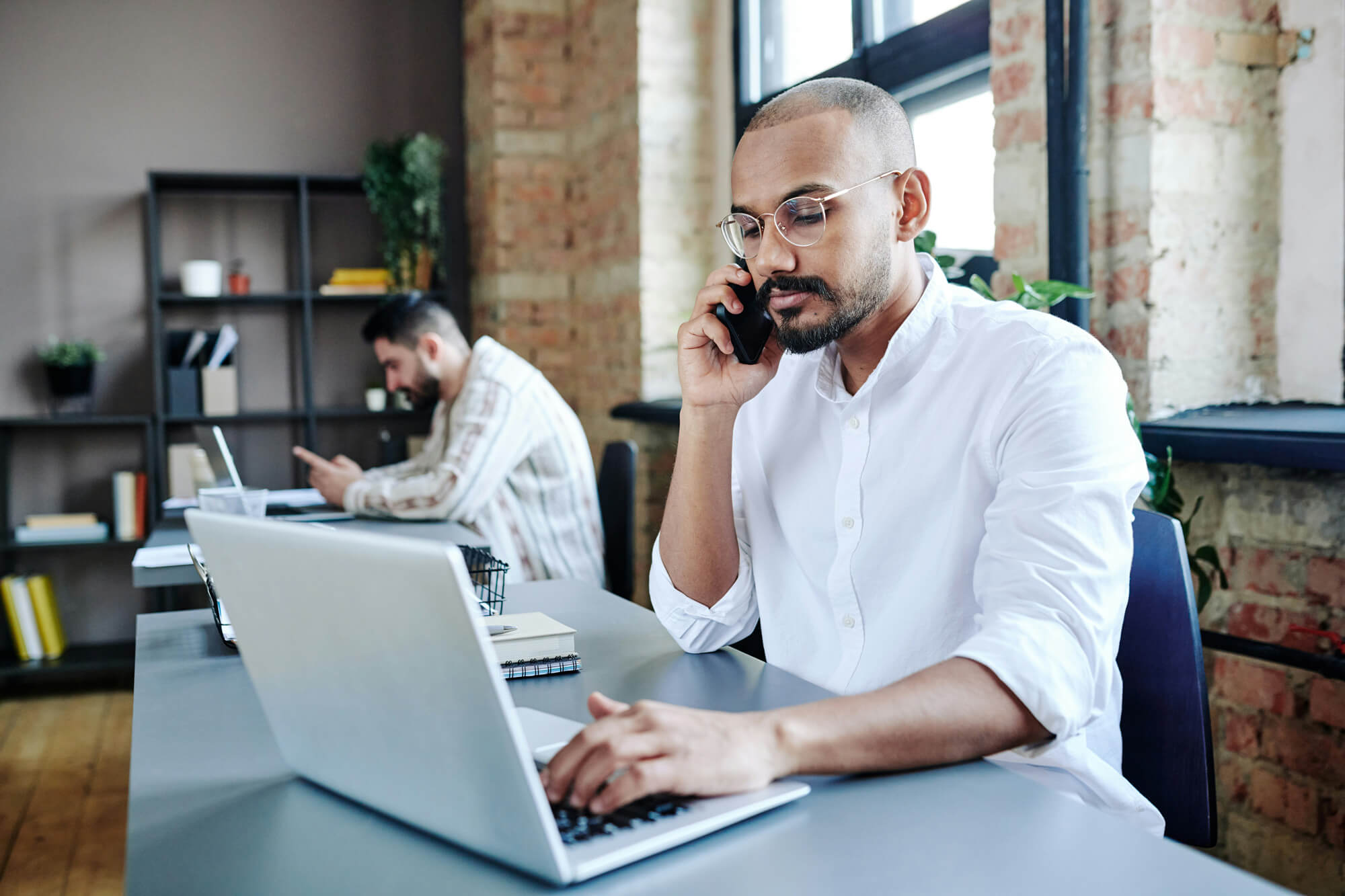 Man in a white shirt and glasses on the phone while working at a laptop in a shared office, a colleague working behind him