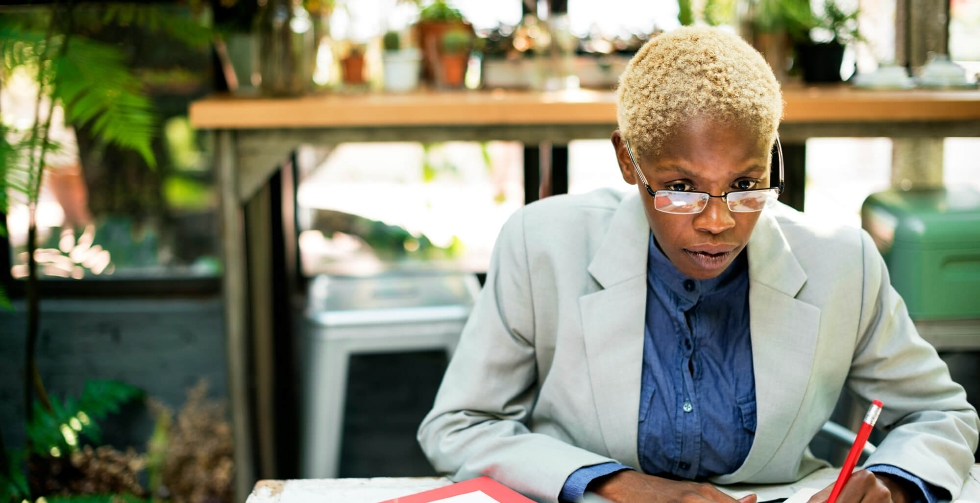 Woman in a gray shirt and clear glasses talking on the phone and gesturing with one hand in a plant-filled office